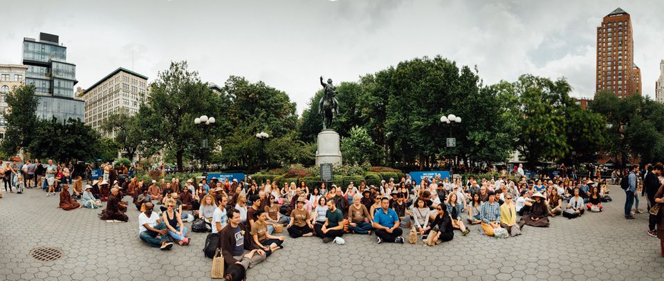 image of people sitting in meditation in a large square in New York City, USA. A park and skyscrapers are visible. 