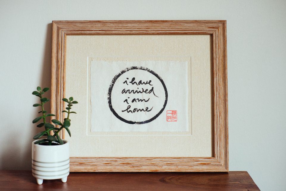 "I have arrived, I am home" calligraphy by Thich Nhat Hanh in a wooden frame, sitting on a wood surface with a small plant