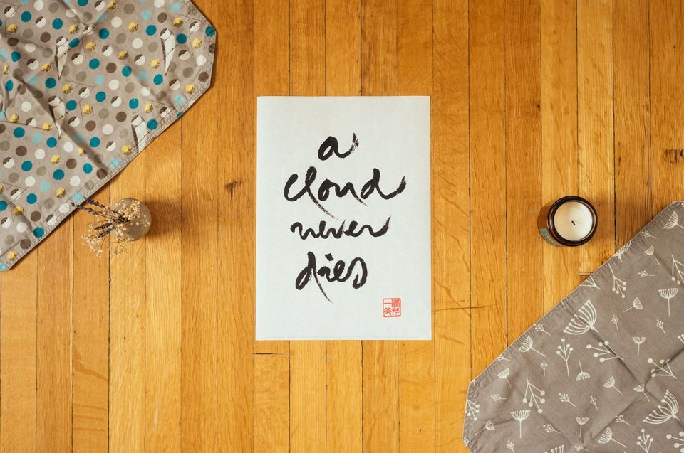 Image of calligraphy, "a cloud never dies." On wooden floor surrounded by two fabric placements, a candle, and dried flowers.